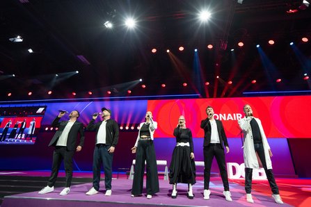 Sechs Personen in schwarz-weißer Kleidung stehen nebeneinander auf der Bühne und singen a cappella in Mikrofone, im Hintergrund bunte Bühnenbeleuchtung und große Leinwand.
