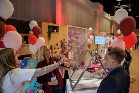 Mehrere Menschen stehen an einem modernen Infostand mit Holzelementen und bunten Ballons. Im Vordergrund sind Losboxen in Pyramidenform zu sehen. Die Personen unterhalten sich angeregt.
