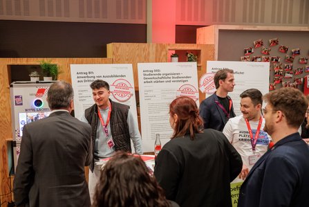 Junge Menschen und Delegierte im Gespräch an einem Ausstellungsstand der DGB-Jugend. Im Hintergrund sind mehrere Plakate mit Antragstiteln und dem Stempel "Noch nicht angenommen" zu sehen. Ein junger Mann lächelt in die Kamera.