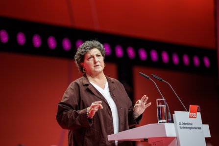 Anja Piel mit kurzen lockigen Haaren steht am Rednerpult und spricht in Richtung Publikum, vor ihr ein Mikrofon und ein Wasserglas, im Hintergrund rote Bühnelemente mit lila Lichtpunkten.
