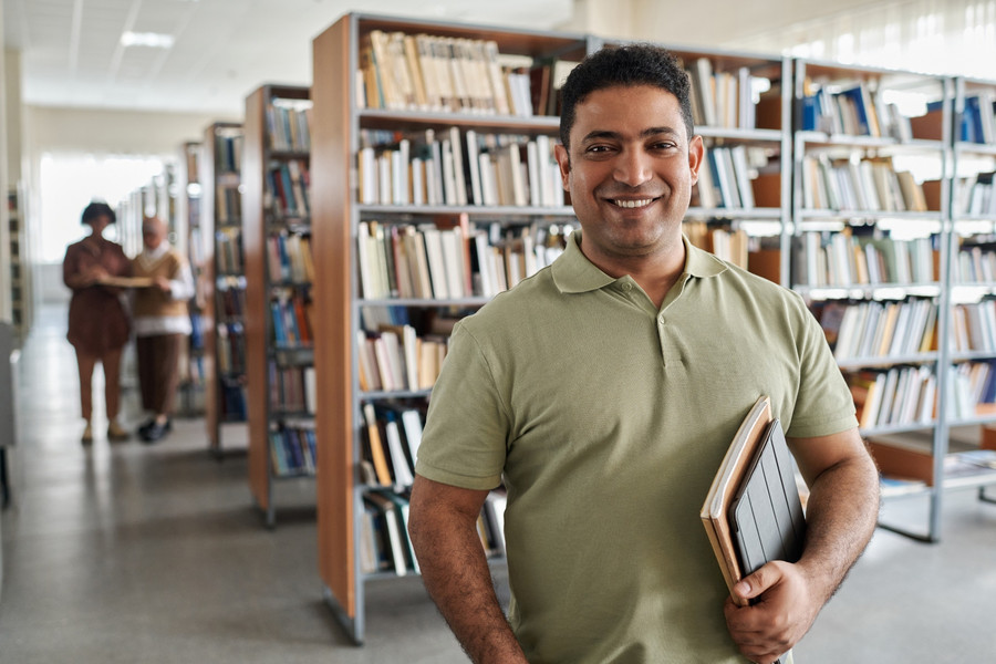 Portrait eines erwachsenen Studenten, der in die Kamera lächelt und in einer Bibliothek steht. 