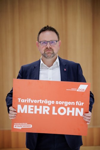 Foto von Guido Zeitler, Vorsitzender der Gewerkschaft NGG, der ein DGB-Plakat in der Hand hält mit der Aufschrift "Tarifverträge sorgen für mehr Lohn" und "Eintreten für die #Tarifwende".