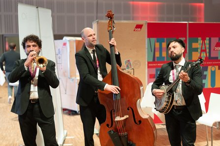 Drei Musiker in dunklen Anzügen spielen live auf einer Ausstellungsfläche. Von links nach rechts: ein Trompeter, ein Kontrabassist und ein Banjospieler. Im Hintergrund sind Messestände zu sehen.