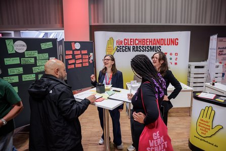 Vier Personen stehen vor einem Stand mit dem Banner „Für Gleichbehandlung – Gegen Rassismus". Zwei Frauen mit roten DGB-Lanyards lächeln, eine hält einen Stift in der Hand. Im Hintergrund sind Pinnwände mit bunten Post-its zu sehen.