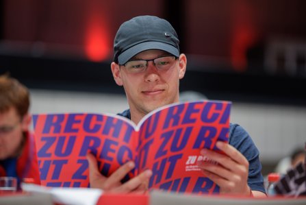 Ein junger Mann mit Brille und dunkler Schildmütze sitzt an einem Tisch und liest lächelnd in einer Broschüre mit dem Titel „Recht auf Rente mit 12 nur Vollzeit". Im Hintergrund unscharf weitere Kongressteilnehmende.