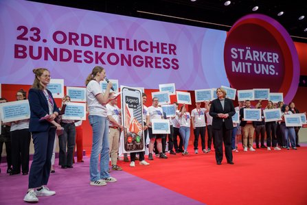 Auf einer Bühne steht eine Gruppe junger Menschen mit Schildern, auf denen Forderungen wie "Die Hochschulen betreiben sexistische Werbung auf dem Arbeitsmarkt" zu lesen sind. Links spricht eine junge Frau am Mikrofon, rechts steht eine Frau im dunklen Blazer. Im Hintergrund ist das Banner "23. Ordentlicher Bundeskongress – Stärker mit uns" zu sehen.