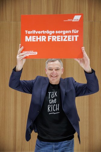 Foto von Jan van Aken, der ein Plakat vom Deutschen Gewerkschaftsbund in den Händen hält mit der  Aufschrift "Tarifverträge sorgen für MEHR FREIZEIT"