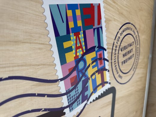 Das Bild zeigt eine Nahaufnahme eines Holzpaneels mit einem bunten Aufkleber, der die Worte "Vielfalt Freiheit" in großen, stilisierten Buchstaben darstellt. Der Aufkleber ist im Design einer Briefmarke gehalten und zeigt lebhafte Farben wie Blau, Rot, Gelb und Violett. Neben dem Aufkleber ist ein runder Stempel zu sehen, auf dem "Vielfalt heißt Freiheit – Eine Ausstellung für Demokratie gegen Ausgrenzung" steht. Das Design ist auffällig und farbenfroh, um das Thema Vielfalt und Freiheit zu betonen.