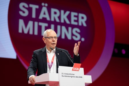 Ein Mann mittleren Alters mit grauen Haaren und Brille steht am Rednerpult und gestikuliert mit der rechten Hand. Er trägt einen schwarzen Anzug mit weißem Hemd. Im Hintergrund ist der Schriftzug "Stärker mit uns" zu sehen.