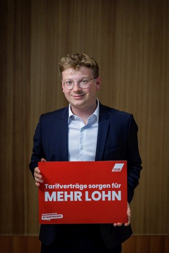 Ben Schneider hält eine rotes Schild mit der Aufschrift "Tarifverträge sorgen für mehr Lohn" im Rahmen der #Tarifwende-Kampagne des DGB
