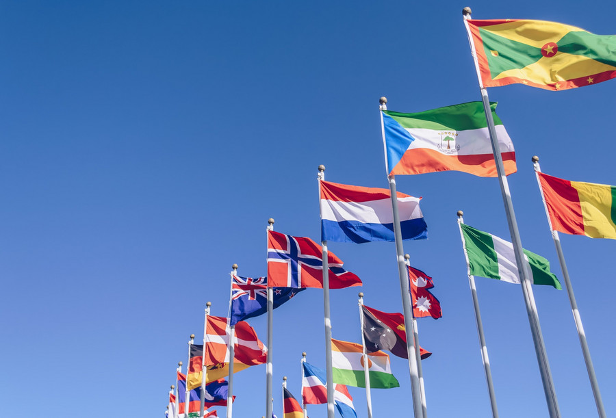 Flaggen verschiedener Länder wehen im Wind mit blauen Himmel im Hintergrund. 