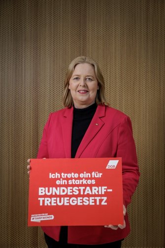 Bärbel Bas (SPD); Bundesministerin für Arbeit und Soziales, hält ein rotes Schild mit der Aufschrift: „Ich trete ein für ein starkes Bundestariftreuegesetz“ und dem Logo des Deutschen Gewerkschaftsbundes (DGB). Darunter steht: „Eintreten für die #Tarifwende“. Der Hintergrund besteht aus einer Holzvertäfelung.