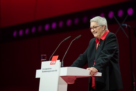 Eine Frau mit kurzem grauen Haar, rotem Kragen und Brille lächelt am Rednerpult, im Hintergrund rote Bühnenelemente mit lila Lichtern und dem Schriftzug "23. Ordentlicher Bundeskongress des DGB".