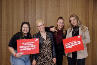 Foto von vier jungen Frauen, die zwei rote Schilder mit der Aufschrift "Ich trete ein für ein starkes Bundestariftreuegesetz" und "Eintreten für die #Tarifwende" halten. Die Schilder stehen für die Unterstützung der #Tarifwende-Kampagne des Deutschen Gewerkschaftsbundes. 