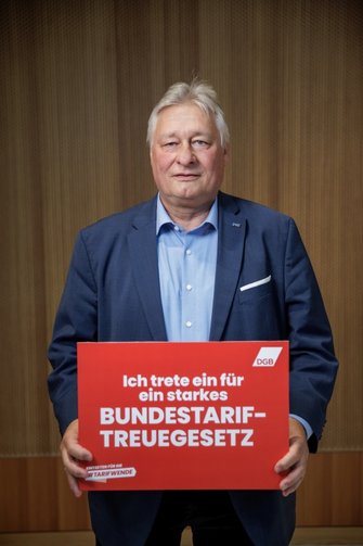 Foto von Martin Burkert, der ein rotes Schild in der Hand hält mit der Aufschrift "Ich trete ein für ein starkes Bundestariftreuegesetz". 