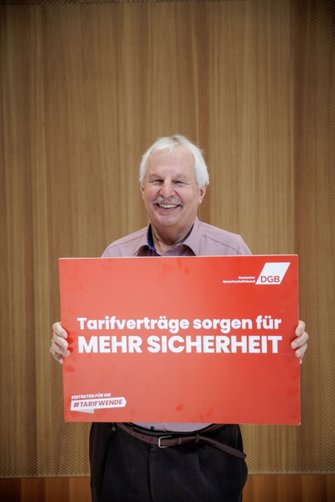 Foto von Horst Kühn, ehemaliger Ingenieur und ver.di-Vertrauensmann, der ein DGB-Plakat in den Händen hält mit der Aufschrift "Tarifverträge sorgen für MEHR SICHERHEIT"