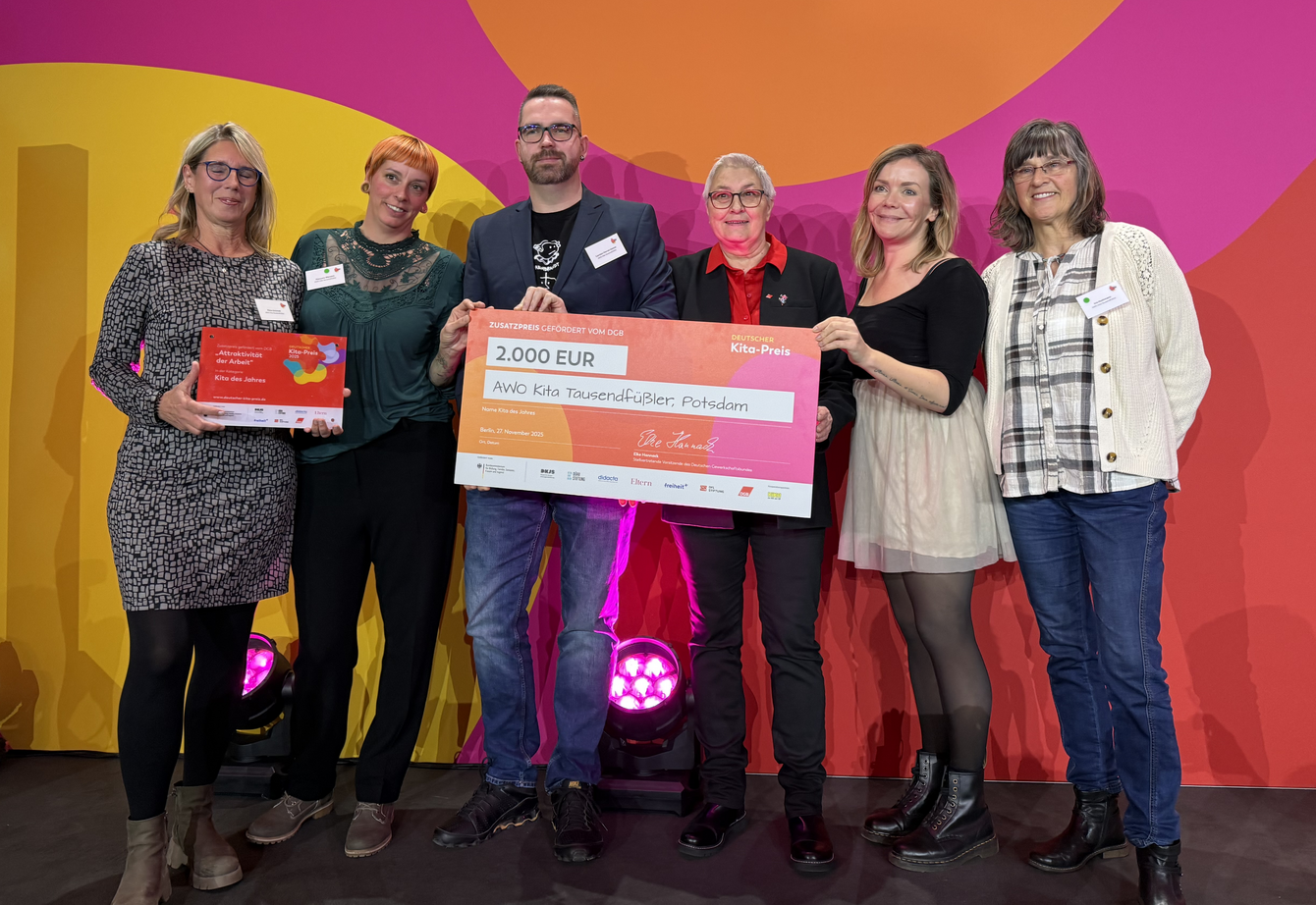 Gruppenbild zeigt 6 Menschen auf einer Bühne, die einen Preis und einen großen Scheck über 2.000 Euro und der Aufschrift "Kita-Preis 2025" und "AWO-Kita Tausendfüßler, Potsdam" halten 