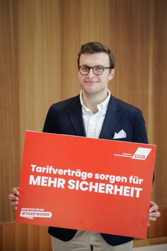 Foto von Phillip-Thomas Kania, Ermittlungsbeamter bei der Finanzkontrolle Schwarzarbeit des Hauptzollamts Gießen, der ein DGB-Plakat in den Händen hält mit der Aufschrift "Tarifverträge sorgen für mehr Sicherheit".