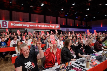 Im großen Kongresssaal halten viele Delegierte rote Stimmkarten hoch, im Hintergrund zeigt ein Banner „Mit Macht für die 8 – Hände weg vom 8-Stunden-Tag!“, rote Sitzreihen und viele Teilnehmende sind zu sehen.