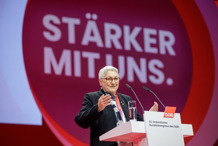 Eine Person mit kurzen grauen Haaren und Brille steht am Rednerpult und gestikuliert mit beiden Händen. Im Hintergrund ist der Schriftzug "STÄRKER MIT UNS." zu sehen.
