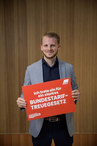 Foto von Tim Klüssendorf, der ein Schild in der Hand hält mit der Aufschrift "Ich trete ein für ein starkes Bundestariftreuegesetz".