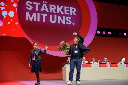 Zwei Personen stehen auf der Bühne des DGB-Bundeskongresses vor dem Slogan „STÄRKER MIT UNS." Die Person rechts hält einen Blumenstrauß und winkt mit erhobener Hand, während die Person links ebenfalls winkt.