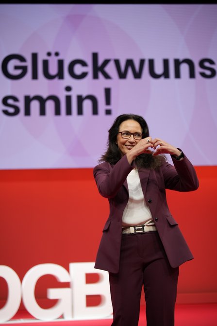 Eine Frau in violettem Hosenanzug und mit Brille steht vor einer Leinwand mit der Aufschrift „Glückwunsch Yasmin!" und formt mit beiden Händen ein Herz vor ihrer Brust. Sie lächelt.