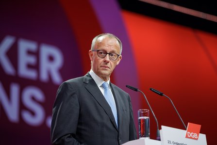 Ein Mann mittleren Alters mit Brille steht am Rednerpult und spricht. Vor ihm zwei Mikrofone und ein Wasserglas. Im Hintergrund verschwommen der Schriftzug "Merz".
