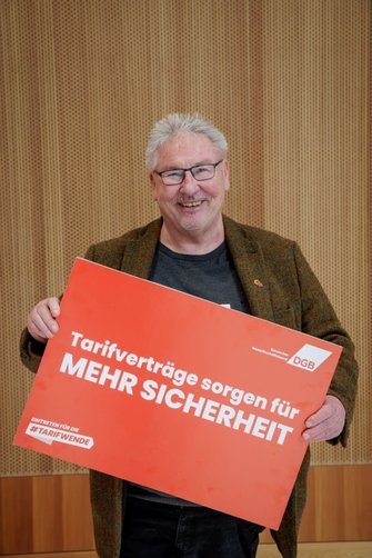 Foto von Peter Witt, der ein rotes Plakat vom DGB in den Händen hält mit der Aufschrift "Tarifverträge sorgen für mehr Sicherheit".